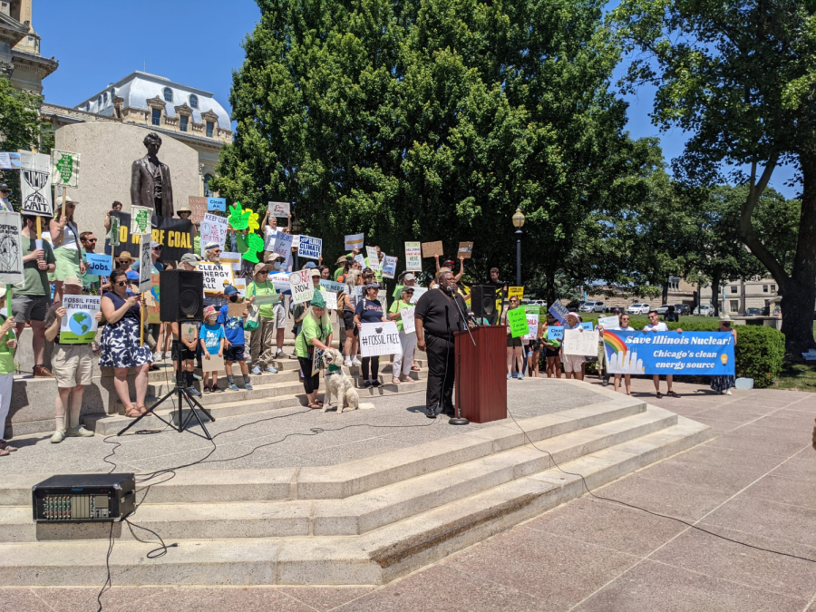 On+steps+and+in+front+of+a+statue%2C+a+large+crowd+mostly+dressed+in+green-and-blue+holds+pro-nuclear+signs.+A+speaker+in+all+black+stands+at+the+podium.