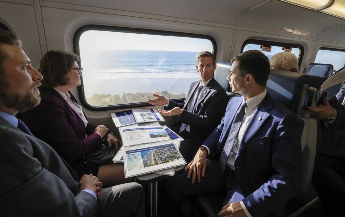 Three men and a woman in suits sit facing each other on a commuter train, ocean waves visible out the window.